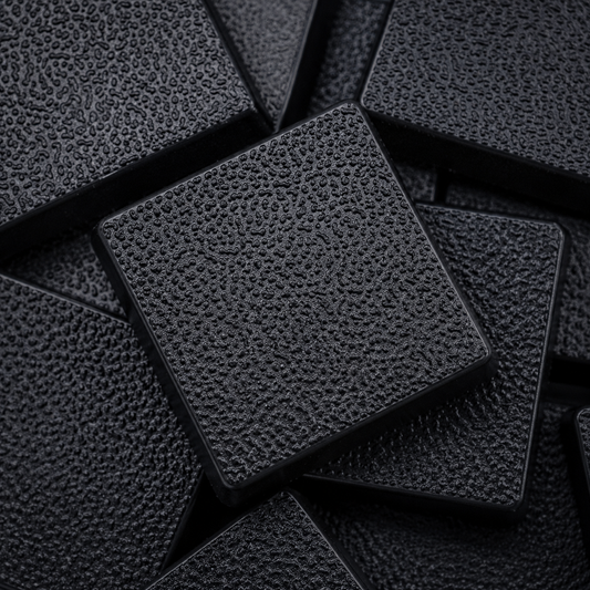 Close-up of black 25mm square plastic miniature bases showing textured top surfaces for tabletop wargaming, RPG figures, rank-and-file units, conversions, and hobby basing projects.